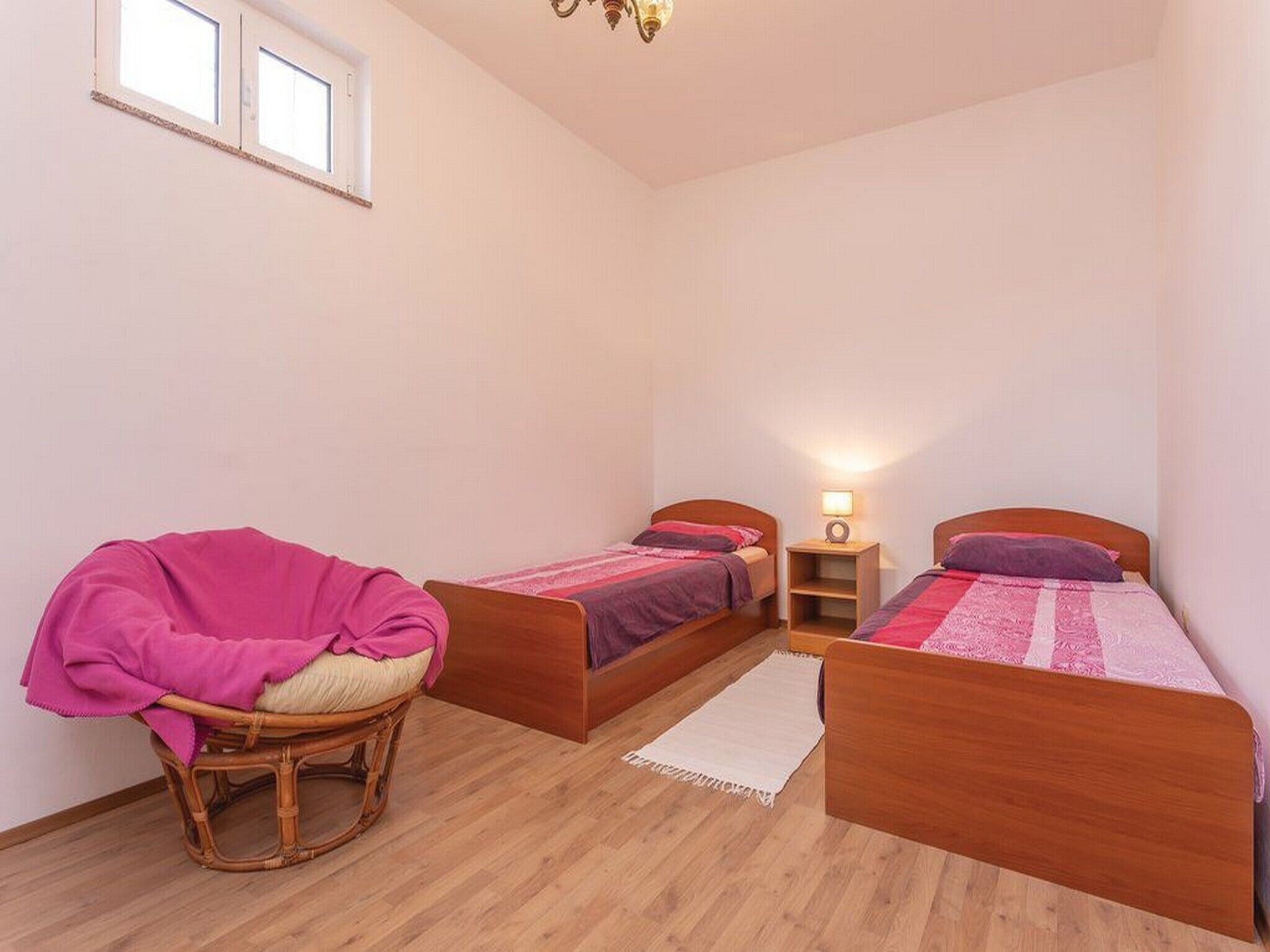 Een slaapkamer met twee houten bedden, een rieten stoel, een klein nachtkastje met een lamp en een raam.