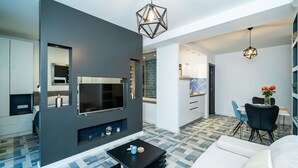 Ein modernes Wohnzimmer mit Flachbildfernseher, einem schwarzen Couchtisch und einer Couch. Im Hintergrund ist eine Küchenzeile mit einem Esstisch und Stühlen zu sehen.