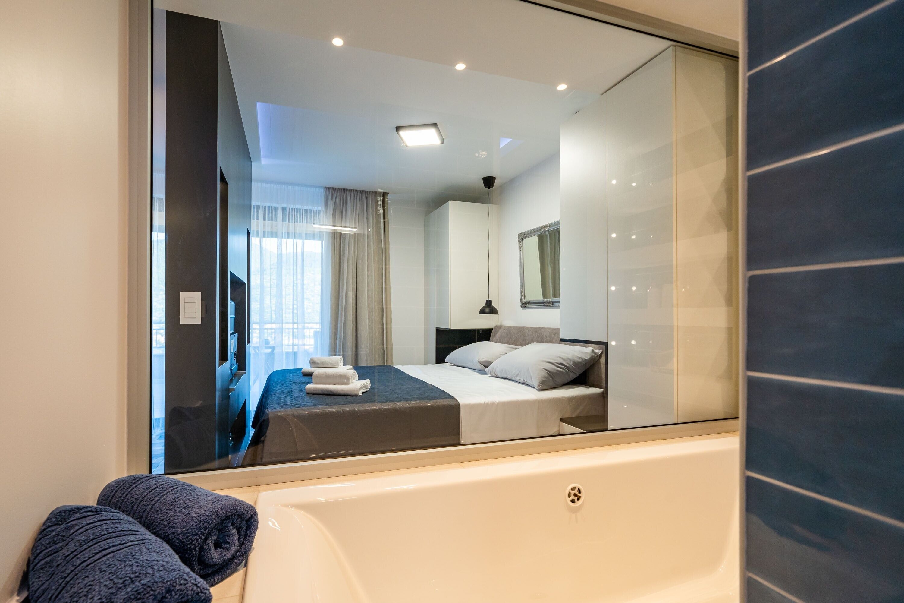 A modern hotel room with a large mirror, a bed, a bathtub, and a view of the city through the window.