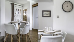 A dining area with a round table and four chairs, a clock on the wall, and a door with curtains. - Port Sunlight, Duke of York Cottage, Grade 2 Listed (wirral)