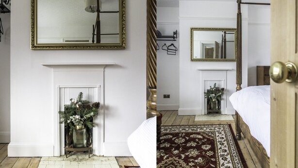 A bedroom with a large bed, a wooden floor, a fireplace, and a mirror with a gold frame. - Port Sunlight, Duke of York Cottage, Grade 2 Listed (wirral)