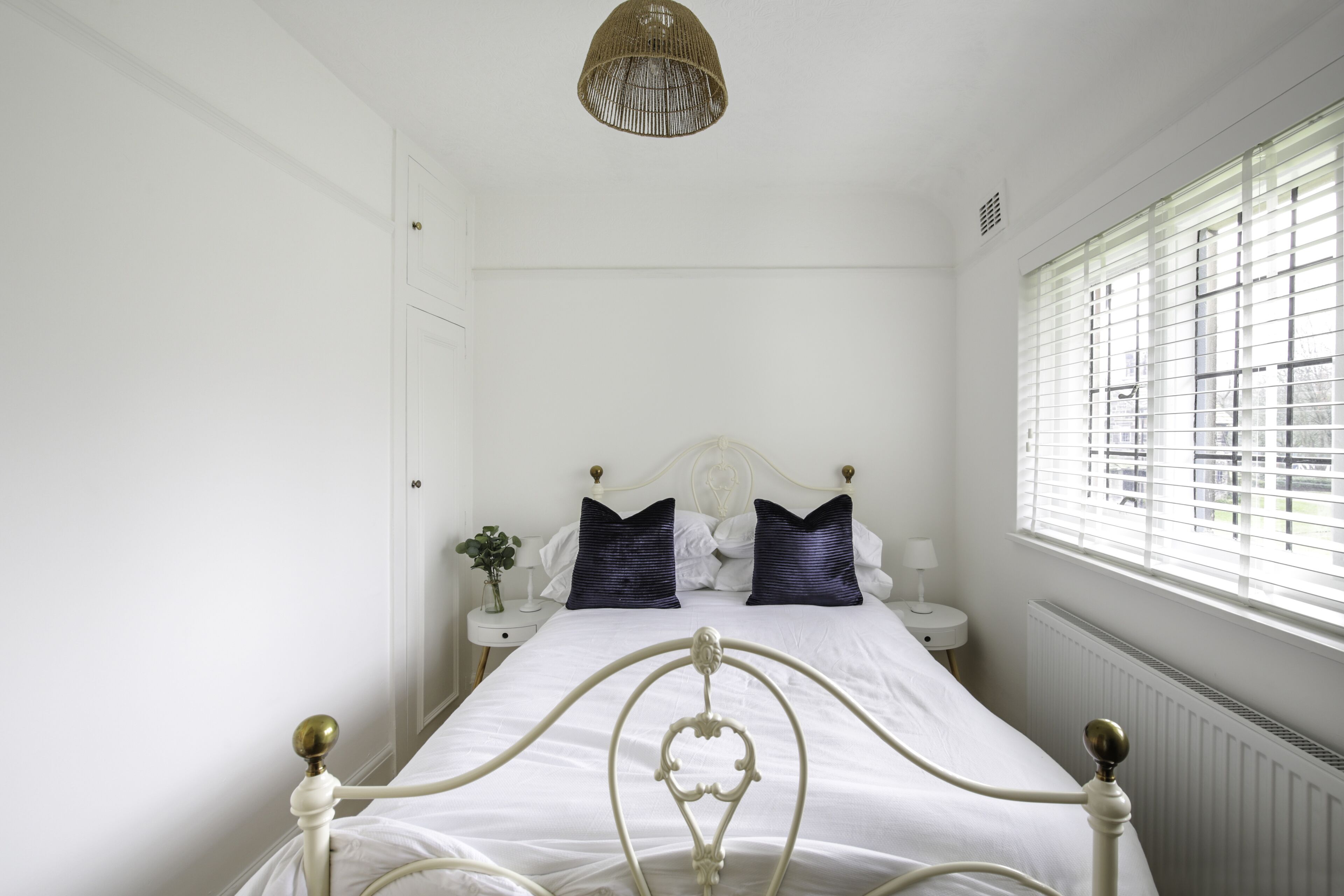 A bedroom with a white bed, two dark pillows, a white nightstand with a lamp, and a window with blinds.