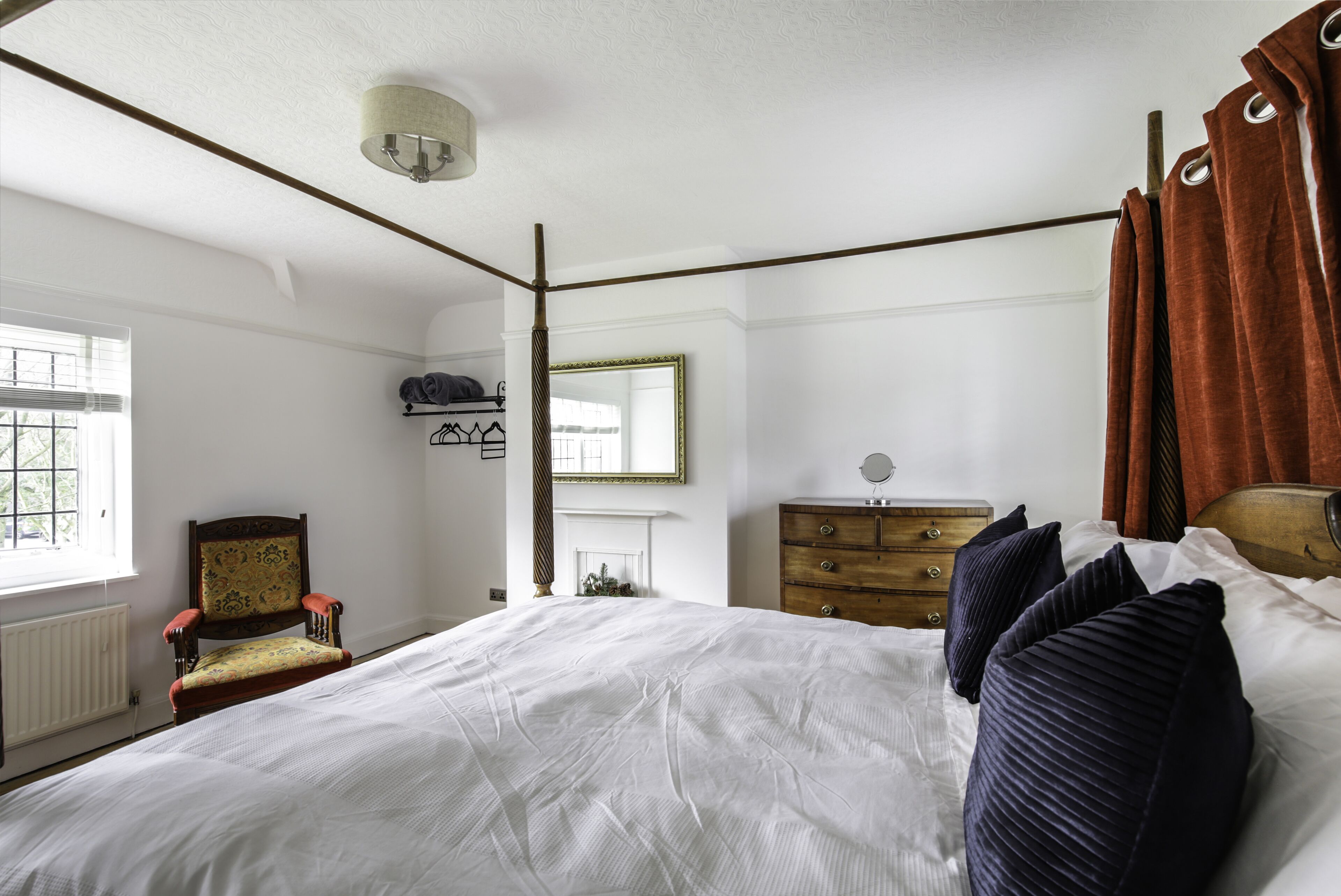 Une chambre à coucher avec un grand lit, une commode en bois, une chaise, un miroir et une fenêtre avec des stores.