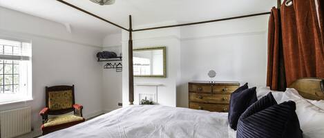 Une chambre à coucher avec un grand lit, une commode en bois, une chaise, un miroir et une fenêtre avec des stores.