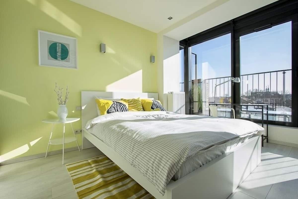A modern bedroom with a large bed, a small side table, a potted plant, and a view of the city through large windows.