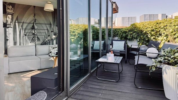 A modern outdoor seating area with a sofa, chairs, and a table, featuring a glass partition and a view of greenery and buildings. - Angel Mint Apartment (Kraków)