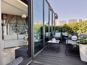 A modern outdoor seating area with a sofa, chairs, and a table, featuring a glass partition and a view of greenery and buildings. - Angel Mint Apartment (Kraków)