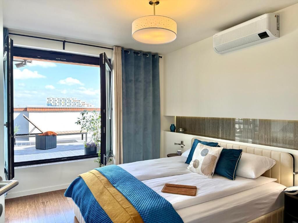 A hotel room with a bed, a bedside table, a window with curtains, a ceiling light fixture, and an air conditioning unit.