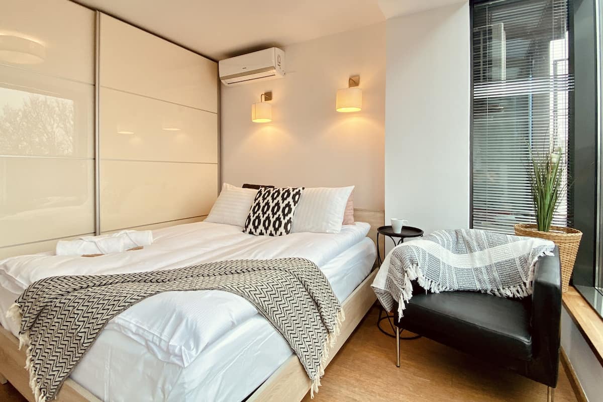 A modern hotel room with a bed, bedside tables, a chair, and a large window with blinds.