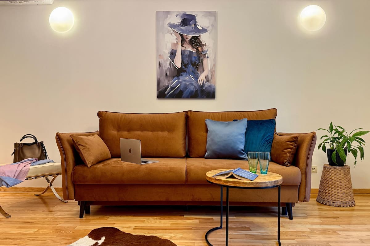 A cozy living room with a brown sofa, a laptop, two glasses, and a book on a side table. There's a cowhide rug, a wicker basket, and a potted plant.