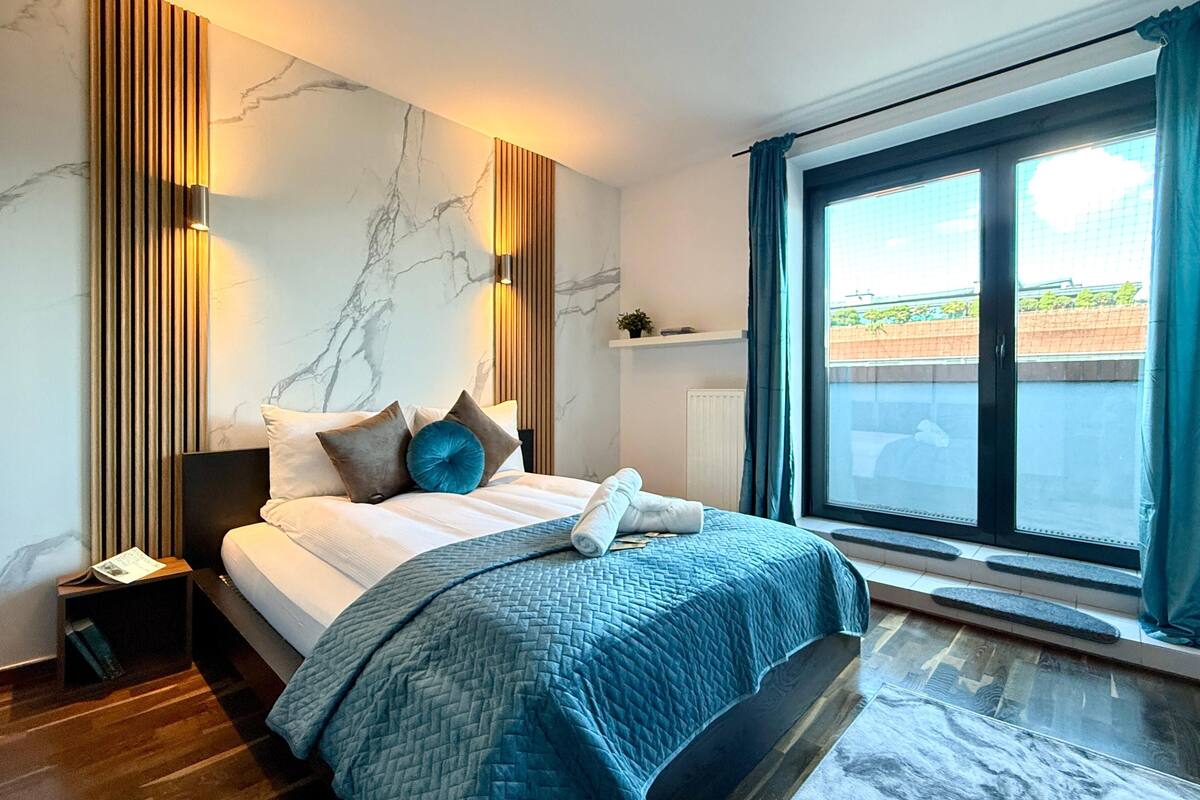 A modern bedroom with a large bed, a nightstand, a wall-mounted light, and a window with curtains.