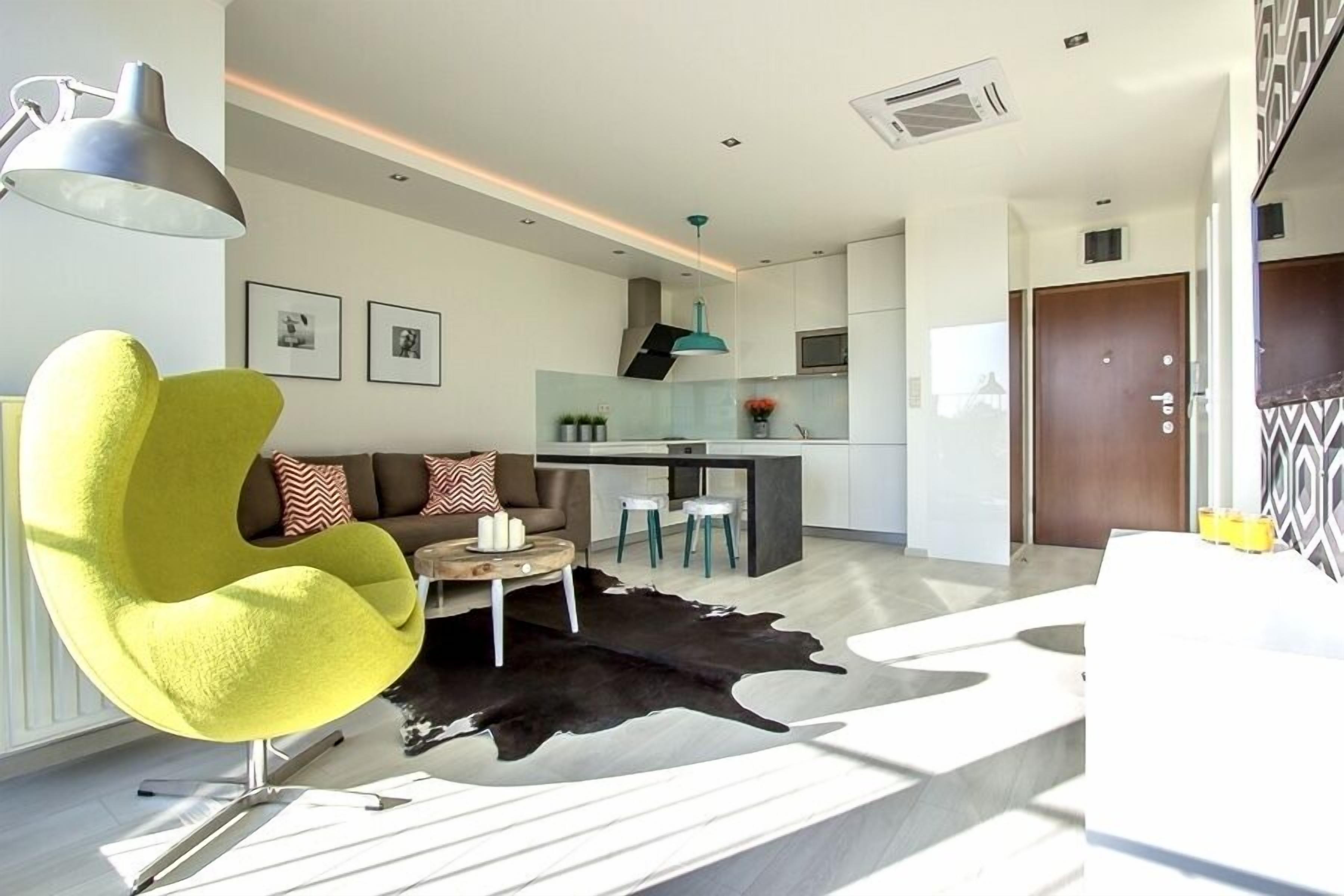 A modern living room with a yellow chair, a sofa with patterned cushions, a coffee table, and a black and white rug.
