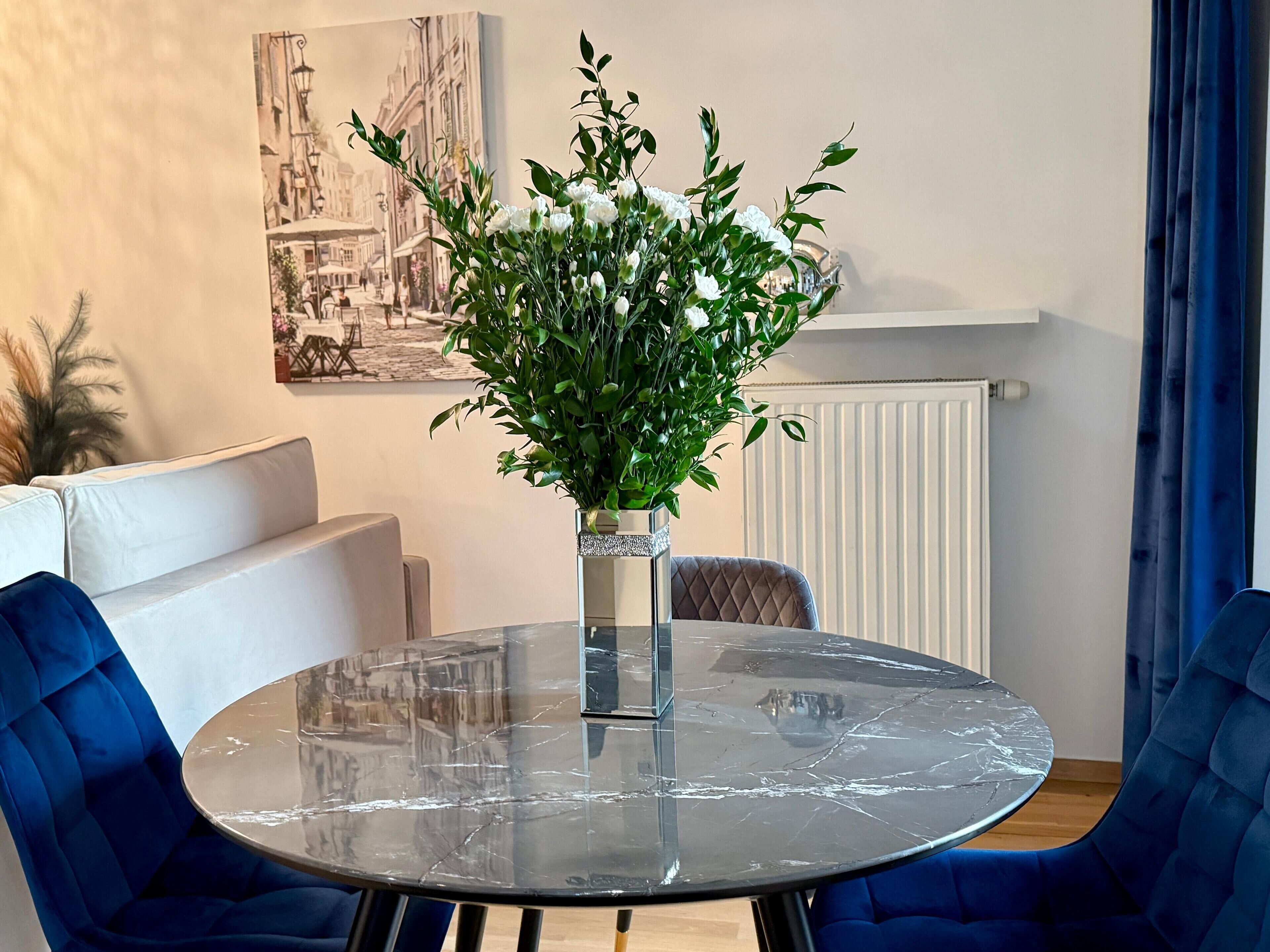 A round glass table with a marble top, surrounded by blue upholstered chairs, a vase with white flowers, and a wall-mounted picture of a street scene.