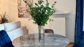 A round glass table with a marble top, surrounded by blue upholstered chairs, a vase with white flowers, and a wall-mounted picture of a street scene.