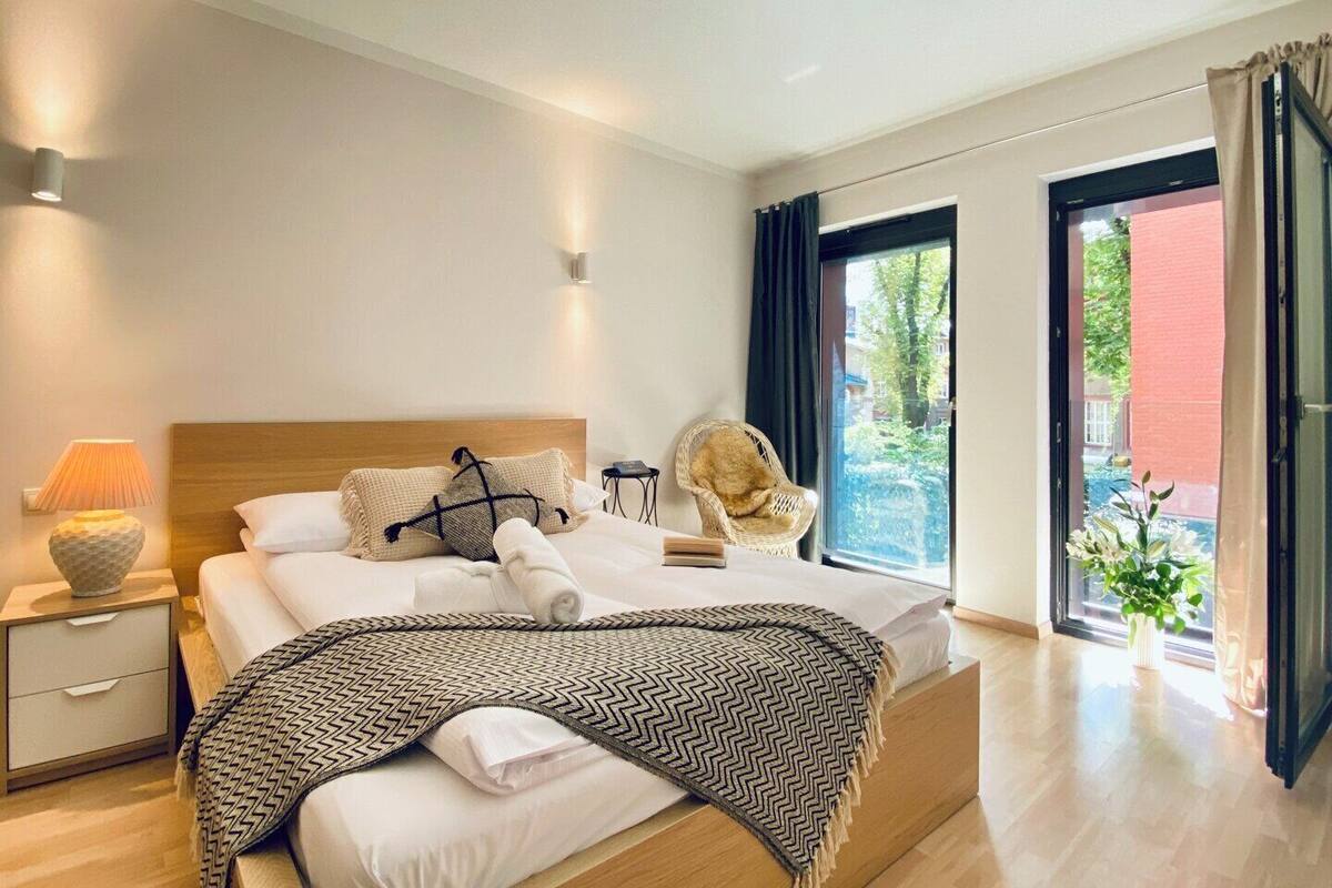 A modern hotel room with a wooden bed, a nightstand with a lamp, a chair, and a view of the outside.