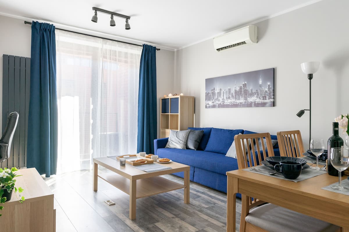 A modern living room with a blue sofa, wooden coffee table, and dining table set with plates and glasses.