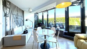 A modern living room with a sofa, dining table, and chairs, large windows with a view of greenery, and a ceiling light fixture. - Angel Mint Apartment (Kraków)