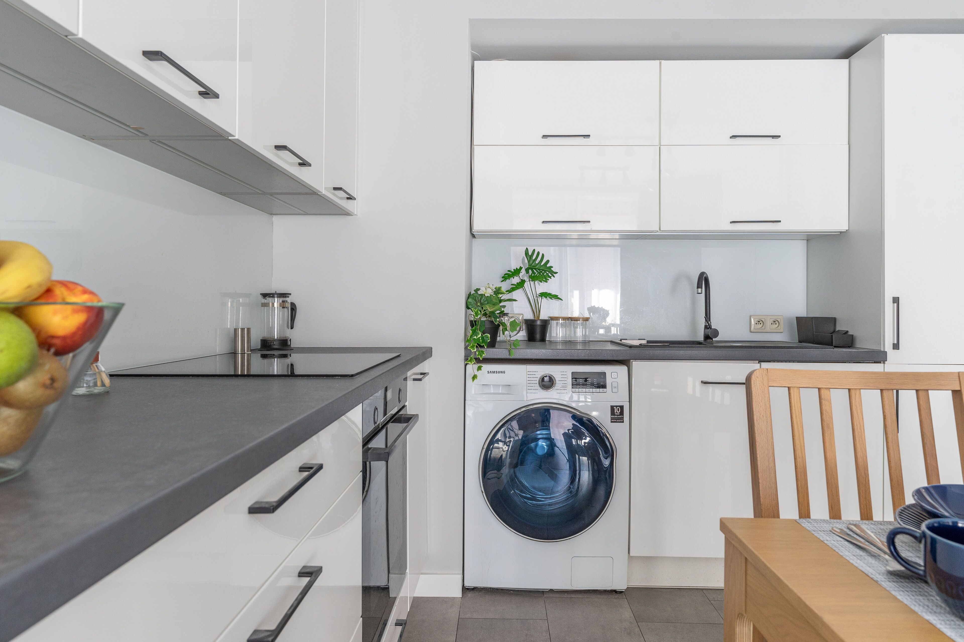 A modern kitchen with white cabinets, a washing machine, a coffee maker, and a bowl of fruit.