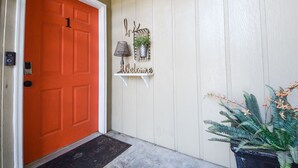 A door with a number 1 sign, a welcome sign, a small shelf with a lamp and a plant, and a potted fern on the porch.