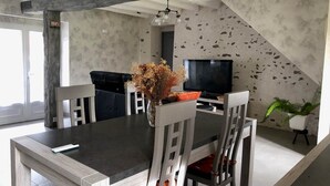 Un comedor con una mesa oscura, sillas blancas, un jarrón con flores secas, un televisor y una planta.