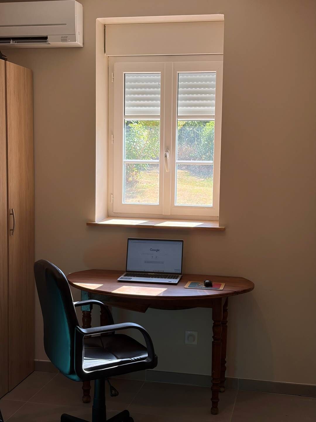Una habitació amb escriptori, portàtil, cadira i finestra amb persianes.