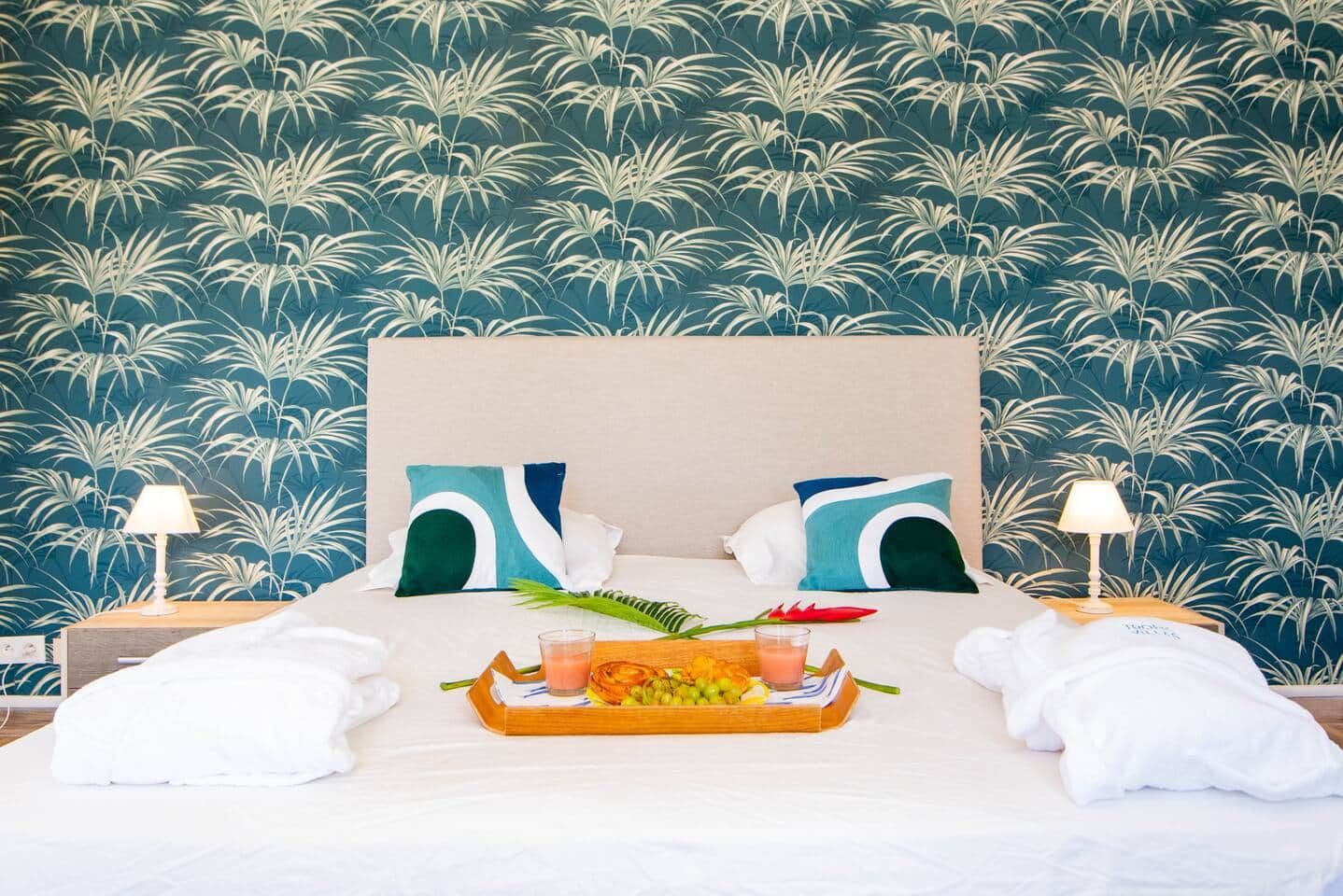 A hotel room with a bed, pillows, a tray with fruits and drinks, and a nightstand with a lamp.