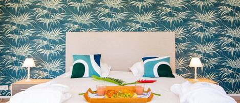 A hotel room with a bed, pillows, a tray with fruits and drinks, and a nightstand with a lamp.