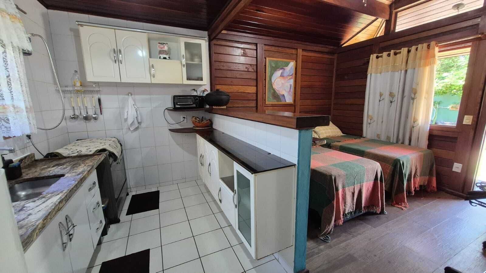 Eine kleine Küche mit weißen Schränken, einer Spüle und einer Arbeitsplatte. Ein Bett mit bunter Decke und ein Fenster mit Vorhängen.