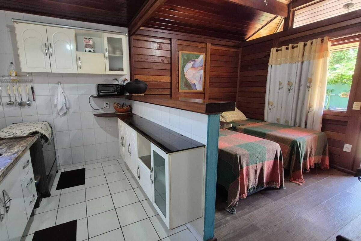 A compact kitchen with white cabinets, a sink, and a small countertop. There is a bed with a colorful blanket and a window with curtains.
