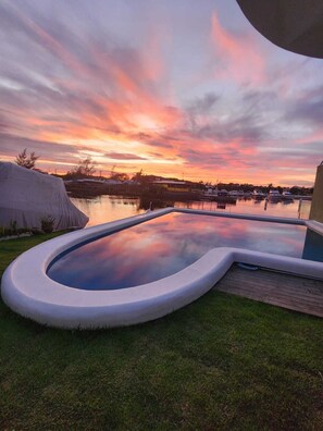A swimming pool with a curved edge, surrounded by grass and a wooden deck. - Morada do Canal Búzios (Búzios)