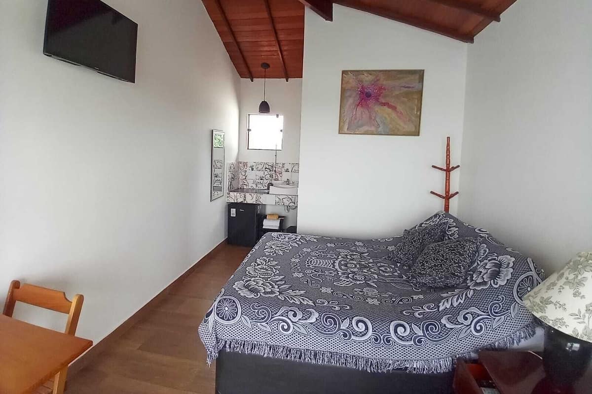 A bedroom with a bed, a desk, a chair, a TV, a painting, and a ceiling fan.