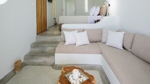 A modern, minimalist living room with a large beige sectional sofa, a wooden coffee table with a candle, and a view of a bedroom with a bed and a TV mounted on the wall. - Located at the breathtaking caldera of Imerovigli (Ημεροβίγλι)