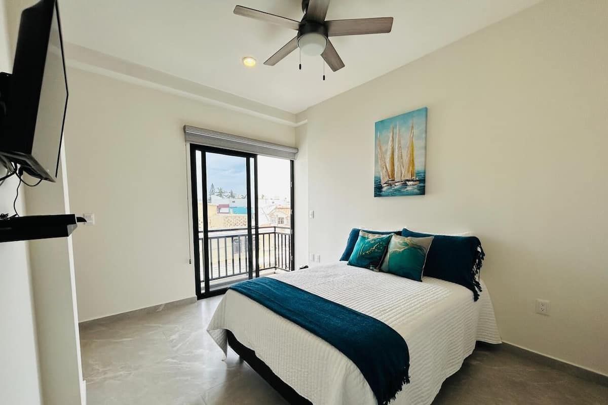 Un dormitorio con cama, ventilador de techo, un cuadro y balcón con vistas.