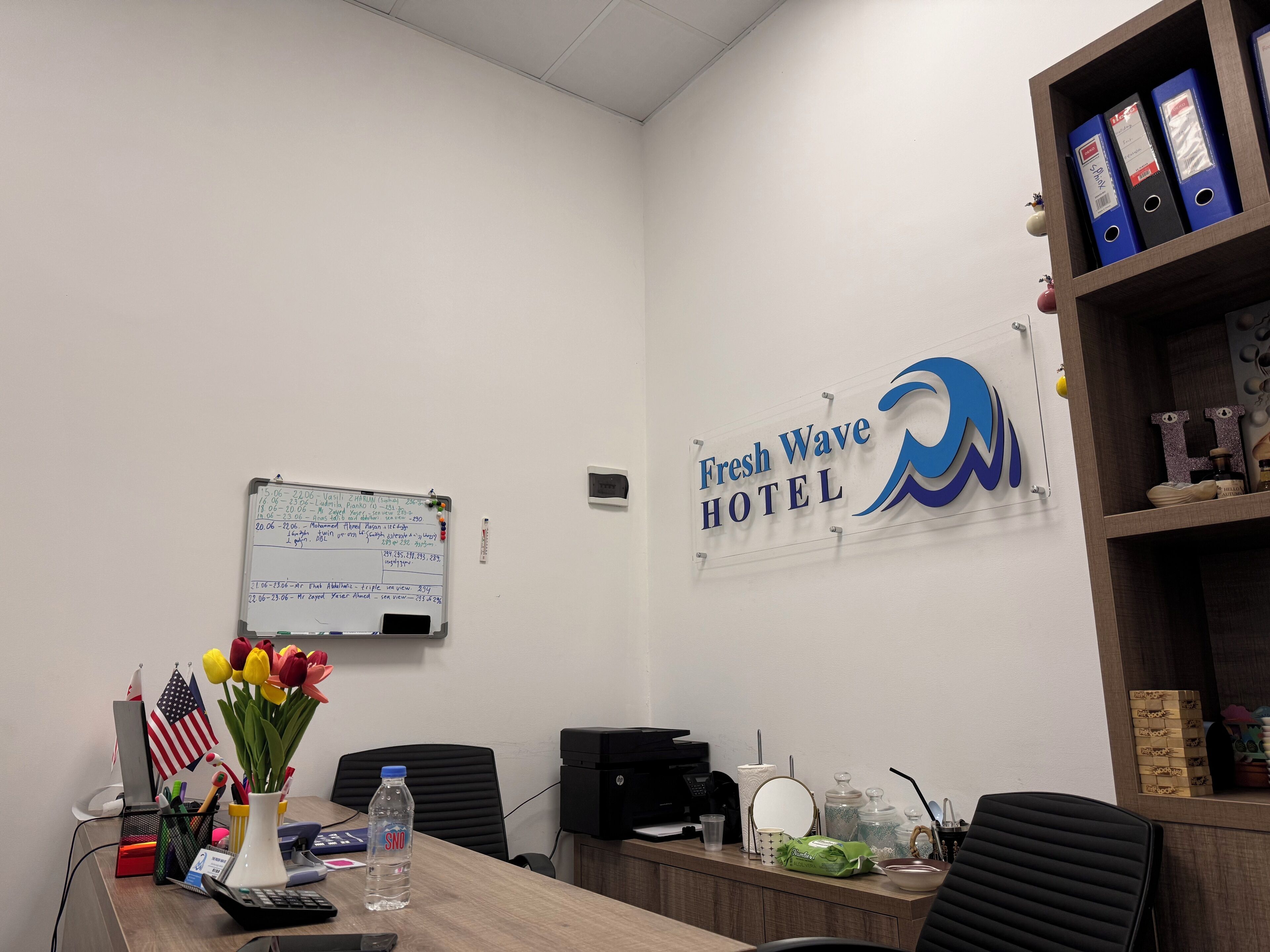 Ein Büro mit einer Whiteboard-Tafel, einem Schreibtisch mit Büromaterialien, einer Wasserflasche und einem Schild für das Fresh Wave Hotel.