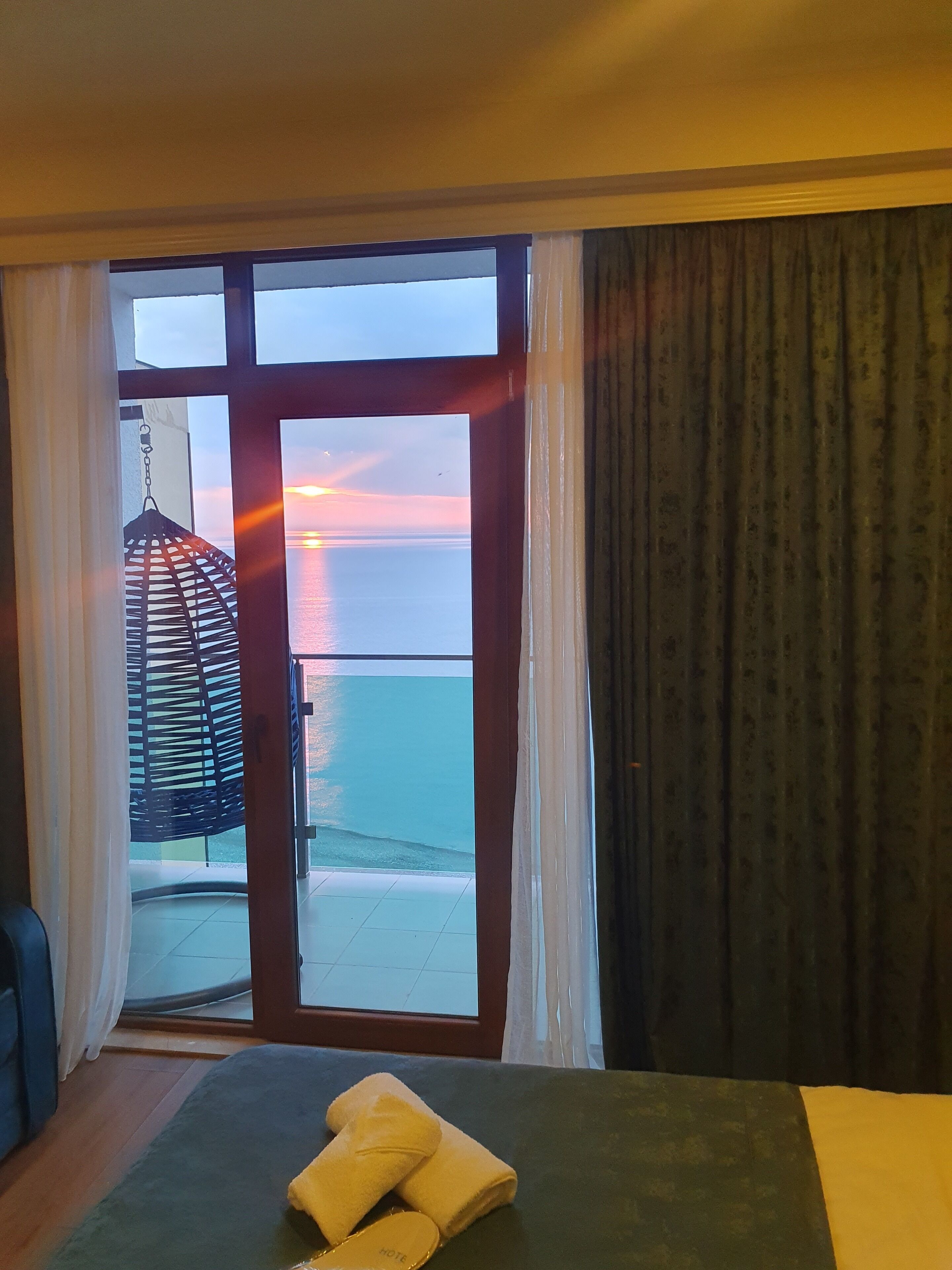 A room with a view of the sunset over the sea, a hanging chair, and a bed with towels.