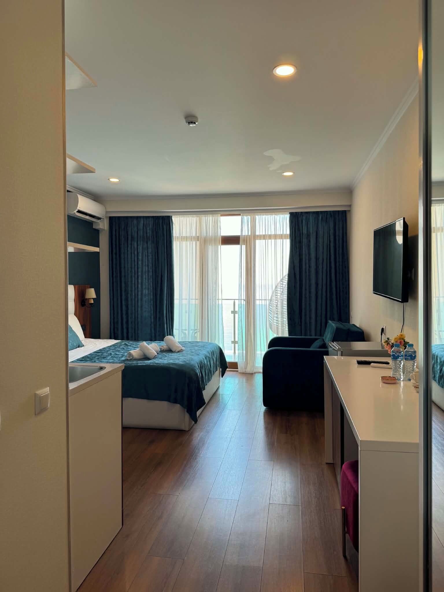 Una habitación de hotel con cama, escritorio, televisor y balcón con cortinas.