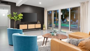 Un salon moderne doté d’un téléviseur à écran plat, de canapés bleus et bruns, d’une table basse en bois et d’une grande porte-fenêtre coulissante donnant sur un espace extérieur avec piscine.