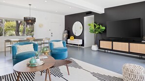 Un salon moderne doté d’un grand téléviseur à écran plat, de fauteuils bleus, d’un coin repas avec une table et des chaises, et d’une grande fenêtre offrant une vue sur les arbres.