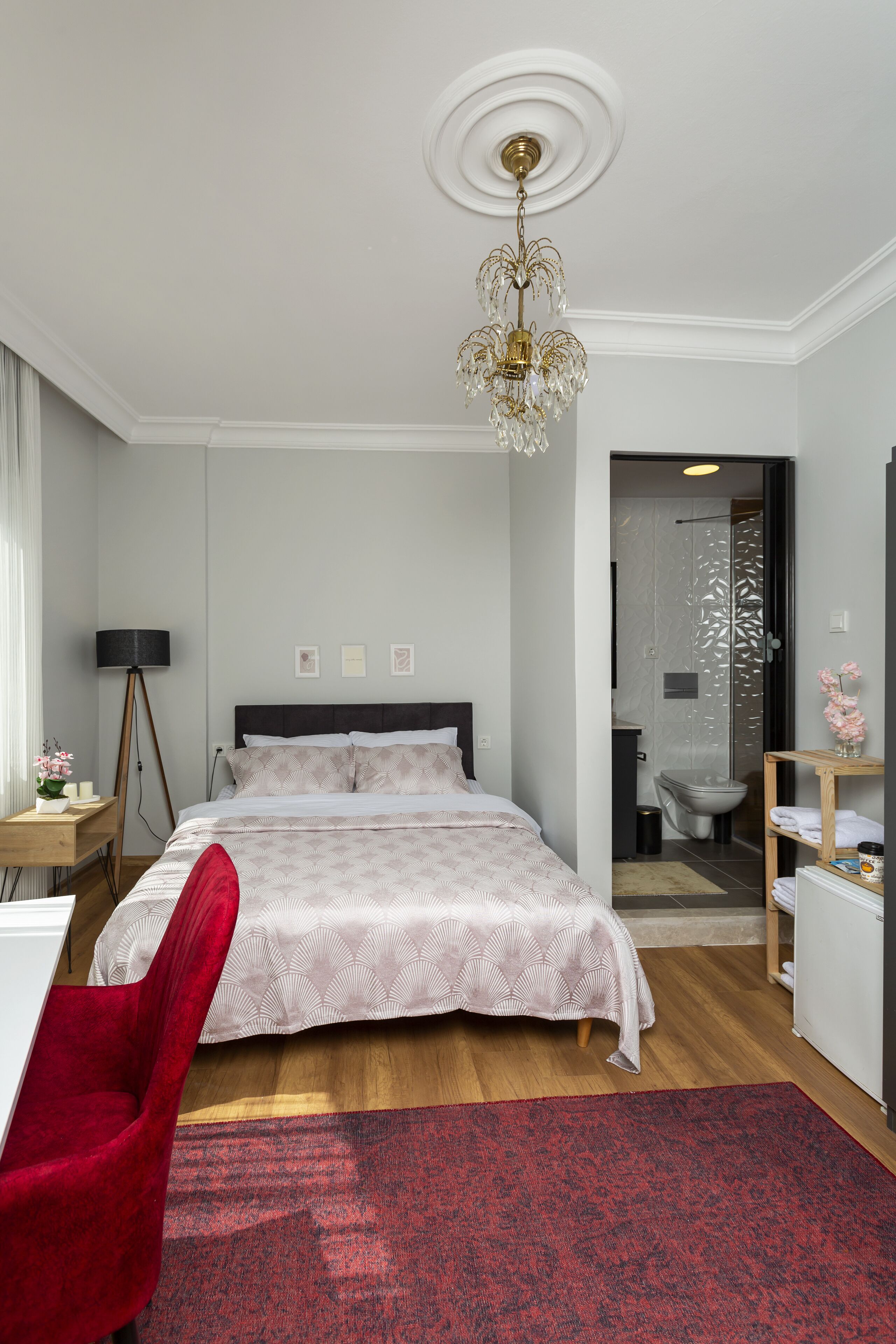 A bedroom with a bed, a red chair, a chandelier, and a bathroom visible through an open door.