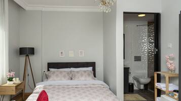 A bedroom with a bed, a red chair, a chandelier, and a bathroom visible through an open door.