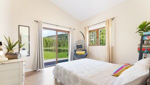 Ein Schlafzimmer mit einem großen Bett, einem Sessel, einem Schreibtisch mit Pflanze und Blick ins Freie.