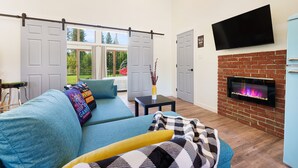 Ein Wohnzimmer mit blauem Sofa, flachbildschirm-Fernseher an der Wand, Kamin aus Ziegelsteinen und einer Glasschiebetür, die zu einem Garten führt.
