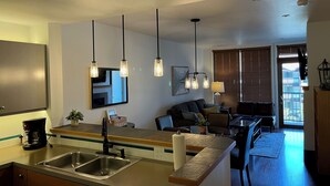 A modern kitchen with a stainless steel sink, a coffee maker, and a dining area with a table and chairs. There is a large window with blinds, a mirror, and pendant lights above the kitchen counter.
