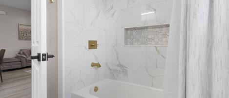 Un baño moderno con pared de mármol, una bañera y una ducha con cabezal dorado.