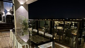 An outdoor dining area with a glass table and white chairs, surrounded by a railing and illuminated by ambient lighting. - Aparamento 2 Dormitorios Edificio Zetta Village Airport (Mariano Roque Alonso)