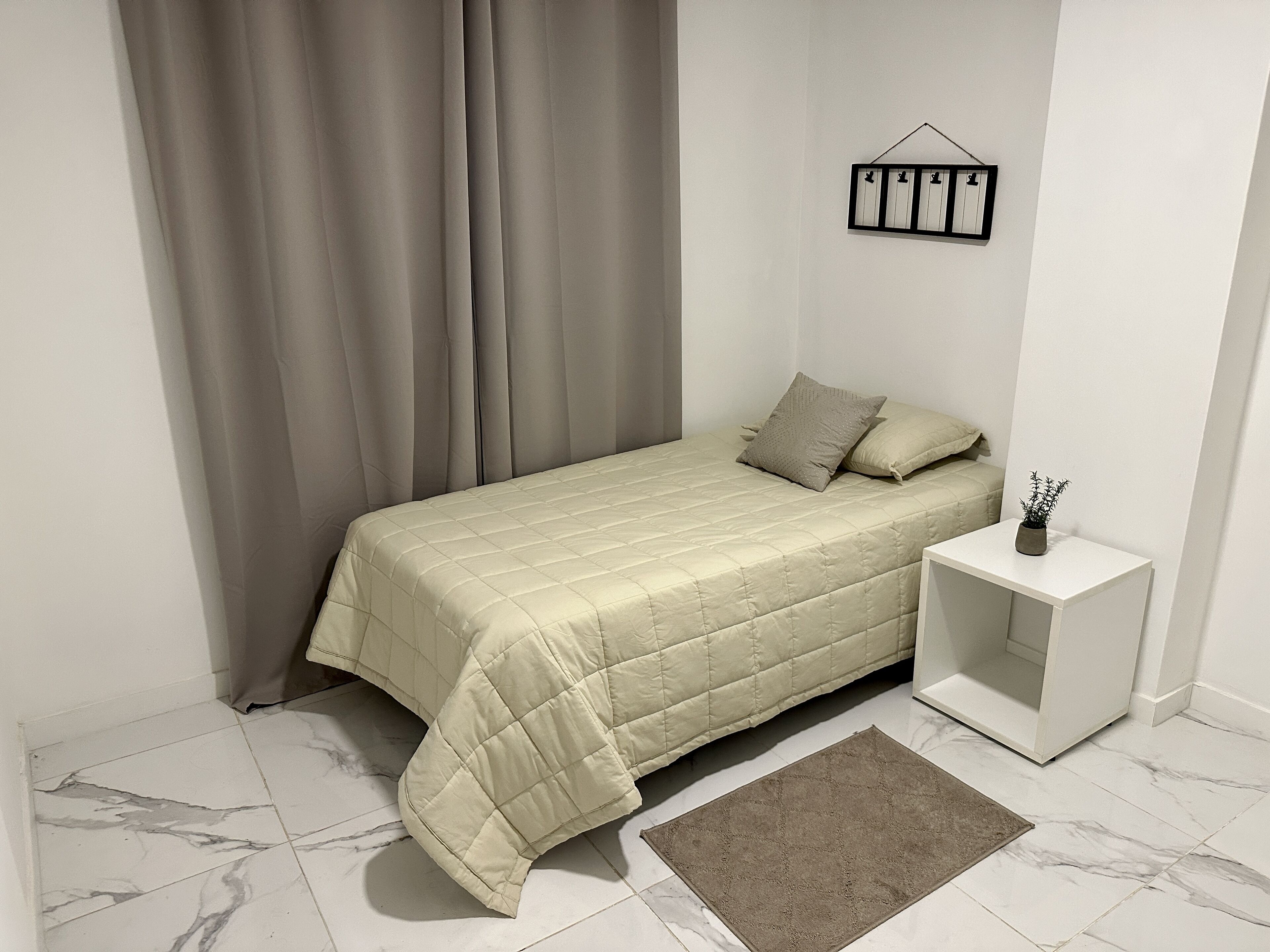 Un dormitorio ordenado con cama, mesita de noche, una pequeña planta en maceta y un marco de fotos colgado en la pared.