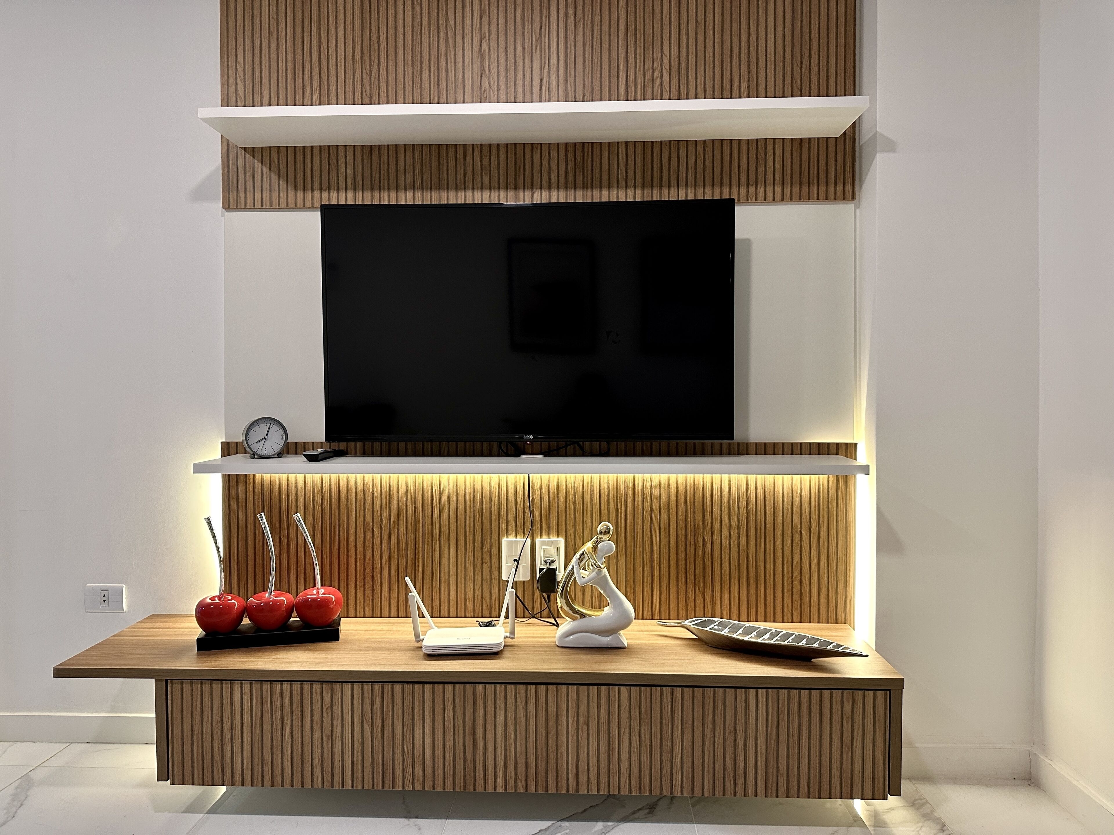 Una sala moderna con un televisor de pantalla plana montado en una pared de madera, un mueble de madera debajo con un router, un jarrón blanco decorativo y un objeto con forma de manzana roja.