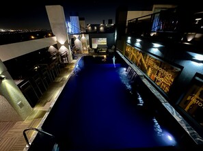 Zona de piscina nocturna con bar y asientos en un lado, y un edificio con señalización iluminada al otro.