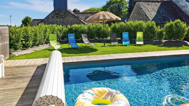 Une piscine avec des chaises longues, une terrasse en bois et un bâtiment en pierre en arrière-plan.