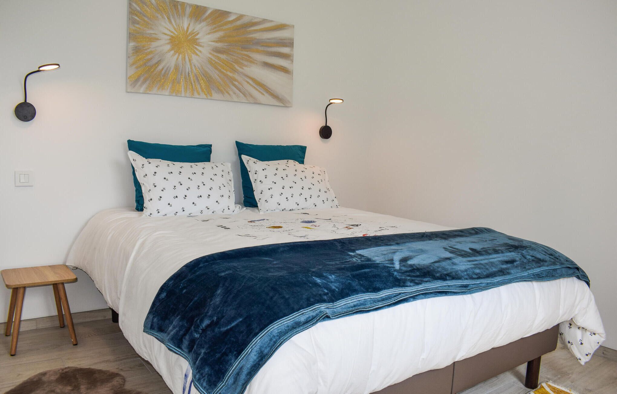 Un lit bien fait, avec une couette blanche et bleue, deux coussins turquoise et un plaid bleu. Deux lampes de lecture sont fixées au mur, de chaque côté du lit.